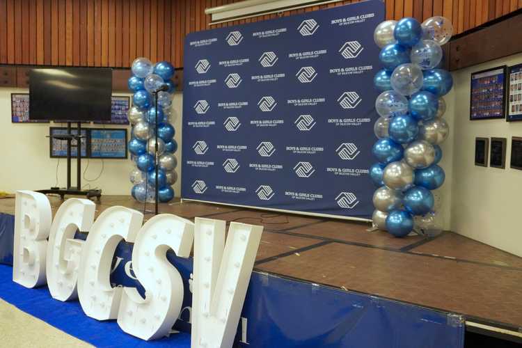 A stage decorated with a blue Boys & Girls Club of Silicon Valley backdrop, balloon stands, and big BGCSV light-up letters.