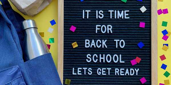 An image from above on a yellow background with confetti. A backpack, water bottle and lunch bag sit beside a letterboard.