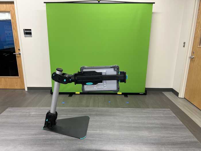 An iPad in a stand facing a green screen. The floor is marked with tape. 