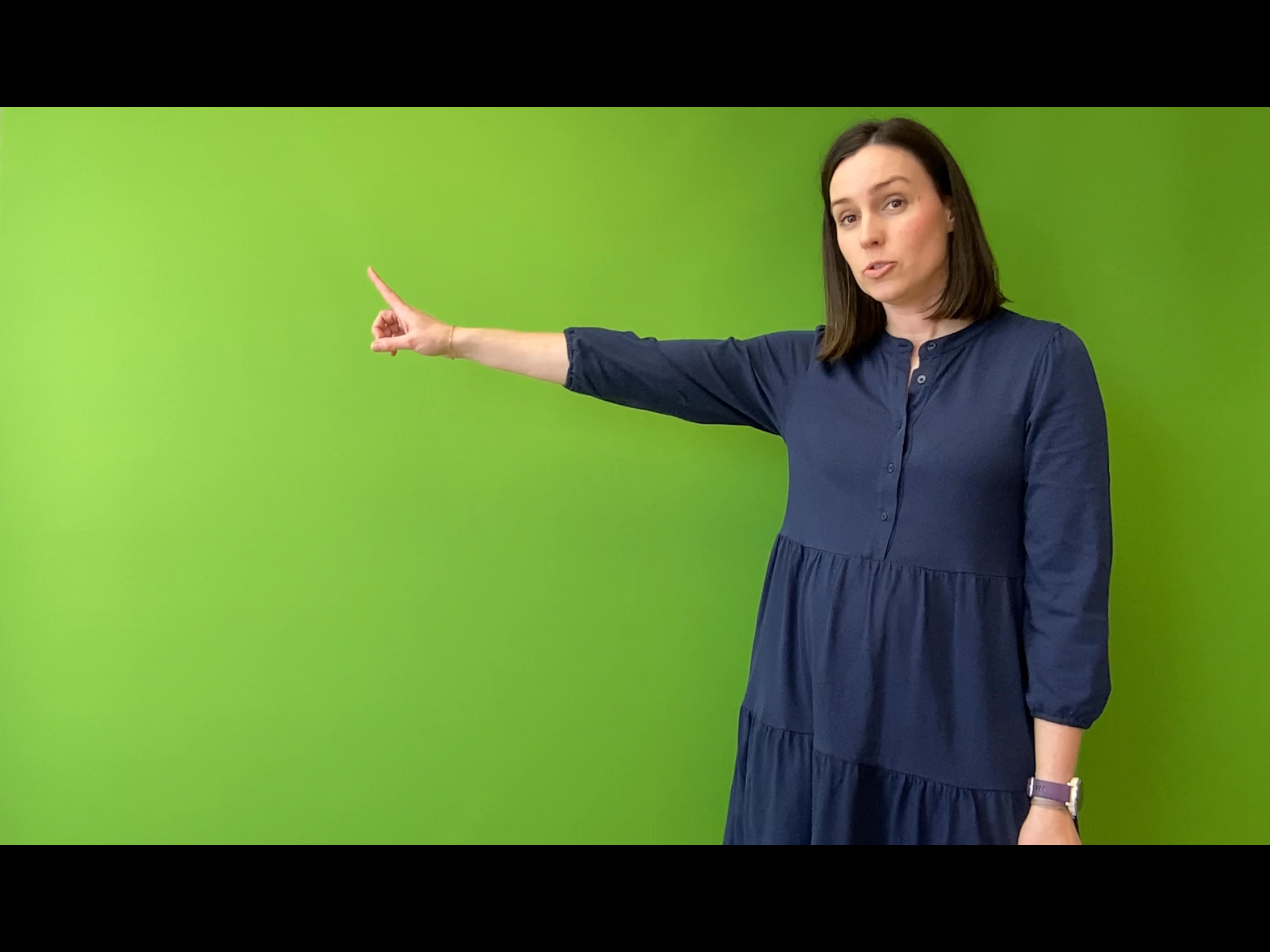 A woman stands in front of a green screen, pointing.